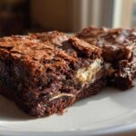 Two fudgy hot chocolate brownies with a shiny, cracked top and visible marshmallow filling on a white plate.