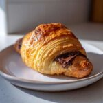 A close-up of a flaky, golden brown chocolate croissant with melted chocolate oozing out, resting on a light plate.