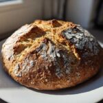 A freshly baked, round loaf of soda bread with a deeply browned, cracked crust dusted with flour, sitting on a white plate.