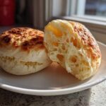 Close-up of two homemade English muffins, one sliced open to show the characteristic airy, open crumb structure.