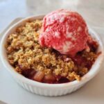 Close-up of a warm rhubarb crisp topped with a scoop of melting strawberry ice cream in a white ramekin.