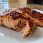 Close-up of two juicy, seasoned air fryer salmon fillets on a white plate, one piece flaked open.