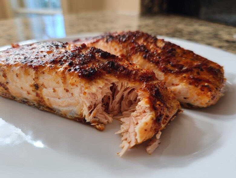 Close-up of two juicy, seasoned air fryer salmon fillets on a white plate, one piece flaked open.