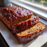 Close-up of a juicy meatloaf loaf with two slices cut, covered in a thick, glossy, dark red glaze.