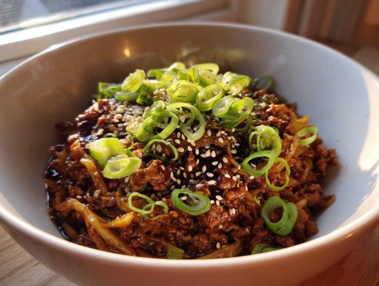 Close-up of a savory keto egg roll in a bowl with ground meat, sauce, topped with sesame seeds and fresh green onions.