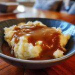 Close-up of creamy mashed potatoes generously covered in smooth, brown chicken gravy in a dark bowl.