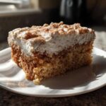 A square slice of moist applesauce cake topped with thick white frosting and dusted with cinnamon.