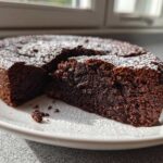 A dark, rich slice cut from a moist chocolate cake dusted with powdered sugar.