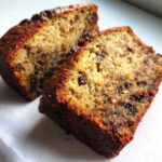 Two close-up slices of moist banana bread studded with melted chocolate chips on a white surface.
