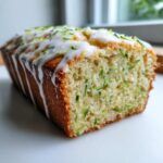 A close-up of a freshly baked loaf of lemon zucchini bread, drizzled with white glaze and topped with zest.