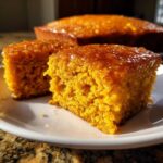 Two squares of vibrant orange sweet potato cornbread topped with a shiny glaze, served on a white plate.