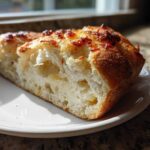 Close-up of a thick slice of airy, no knead bread with a golden, slightly charred topping, served on a white plate.