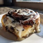 A single, decadent Oreo cinnamon rolls, drizzled with white icing and topped with crushed chocolate cookies, sitting on a white plate.
