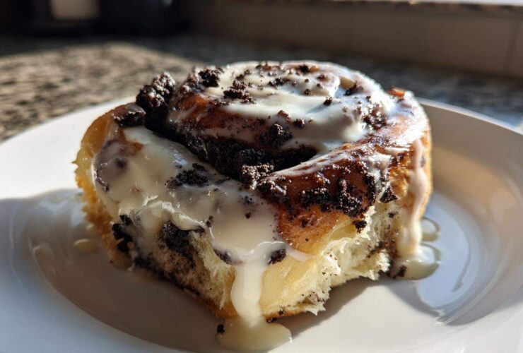 A single, decadent Oreo cinnamon rolls, drizzled with white icing and topped with crushed chocolate cookies, sitting on a white plate.