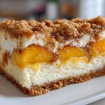 Close-up of a rich slice of peach cobbler cheesecake featuring layers of creamy filling, bright peaches, and a brown sugar crumble topping.