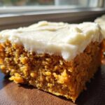 A close-up shot of a moist, orange-colored slice of pumpkin bars with thick, white cream cheese frosting.