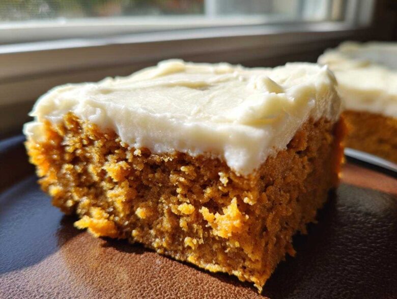 A close-up shot of a moist, orange-colored slice of pumpkin bars with thick, white cream cheese frosting.