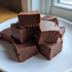 A stack of rich, dark chocolate fudge squares piled on a white plate near a bright window.