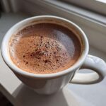 Close-up of rich, foamy mexican hot chocolate served in a white mug on a sunlit windowsill.