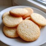 A close-up of several round, golden-edged sugar cookies piled on a white plate near a window.