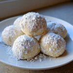 A close-up of several delicious Russian tea cakes heavily coated in powdered sugar, piled on a white plate.