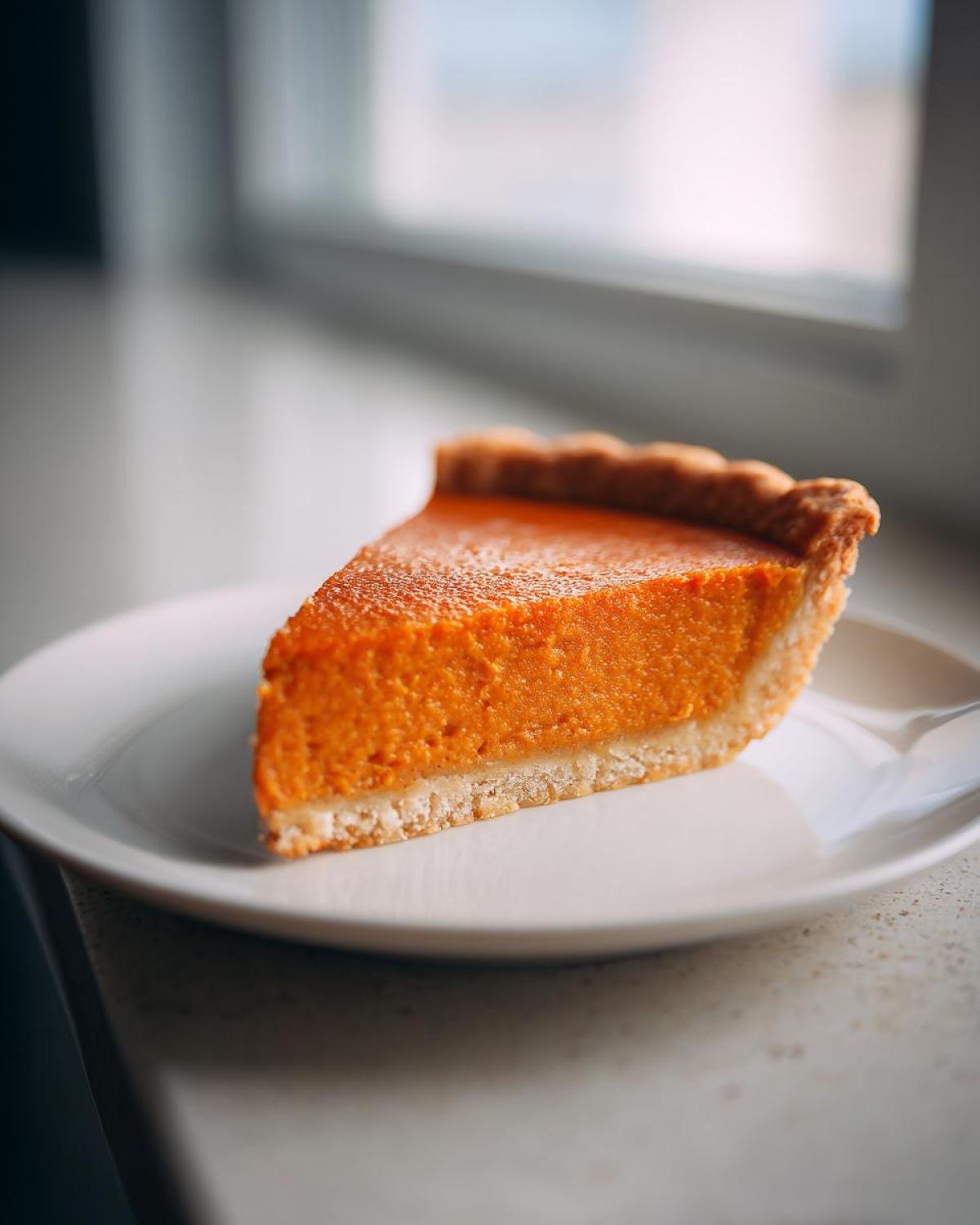 A close-up side view of a perfectly baked slice of easy pumpkin pie resting on a white plate near a bright window.