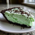Close-up of a thick slice of light green grasshopper pie with a dark chocolate crust, topped with chocolate crumbs.