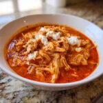 A close-up of a white bowl filled with shredded slow cooker buffalo chicken chili, topped with dollops of white cheese.