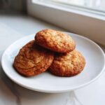 Three soft, round applesauce cookies coated in cinnamon sugar stacked on a white plate.
