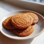 Four soft, crinkled applesauce cookies stacked slightly on a small, light grey plate near a window.