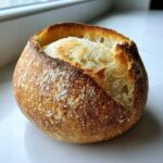 A close-up of a rustic, round sourdough bread bowl with a deeply browned, flour-dusted crust and an open top.