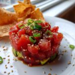 A vibrant stack of spicy tuna tartare layered over avocado, garnished with sesame seeds and scallions, served with crispy wonton chips.