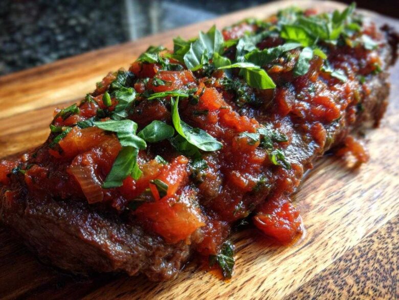 Close-up of a juicy steak pizzaiola covered in rich tomato sauce and fresh basil leaves.