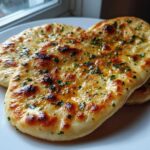 Two pieces of fluffy homemade naan bread, brushed with butter and sprinkled with green herbs, resting on a white plate.