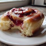 A close-up of a soft, glazed Strawberry Cheesecake Sweet Rolls with a bite taken out, showing the creamy filling.