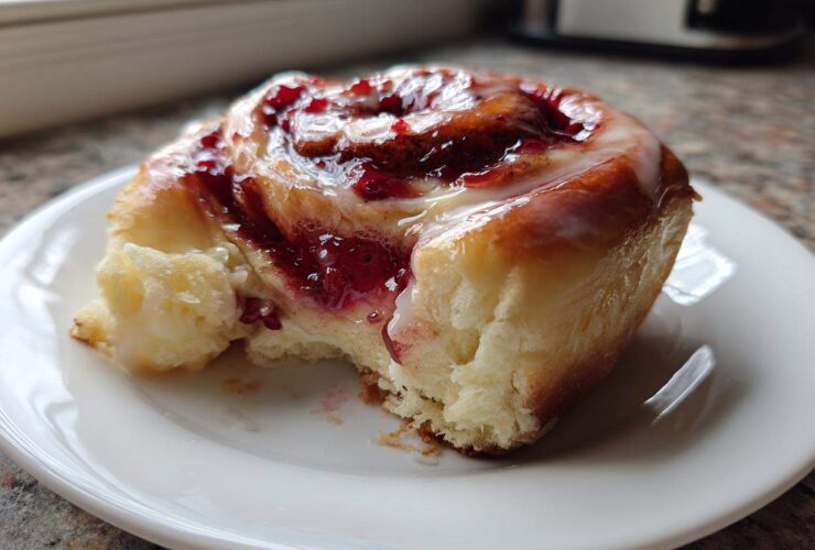 A close-up of a soft, glazed Strawberry Cheesecake Sweet Rolls with a bite taken out, showing the creamy filling.