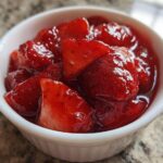 Close-up of chunky strawberry freezer jam glistening in a small white bowl on a granite countertop.