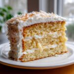 A tall, moist slice of 3-layer coconut cake with white frosting and toasted coconut flakes on top.