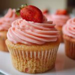 A close-up of a freshly baked strawberry cupcake topped with pink frosting and a fresh strawberry.