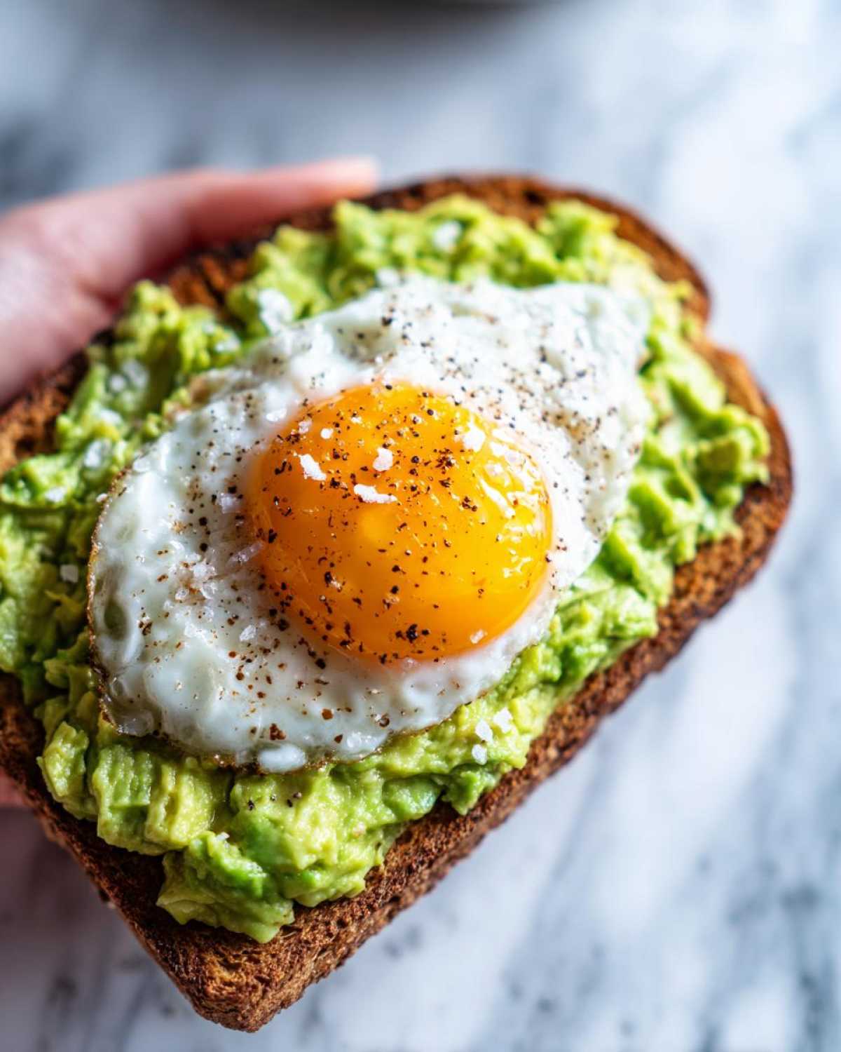 A close-up of delicious avocado toast topped with a sunny-side-up fried egg and cracked pepper.