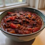 Close-up of a steaming bowl of rich, thick black bean chili topped with chunks of tomato.