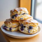 A stack of three freshly baked blueberry scones topped with a sweet white glaze, sitting on a light plate.