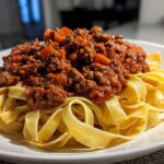Close-up of rich, meaty bolognese sauce generously spooned over a bed of fresh tagliatelle pasta on a white plate.