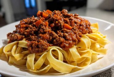 Close-up of rich, meaty bolognese sauce generously spooned over a bed of fresh tagliatelle pasta on a white plate.