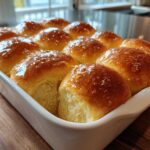 A white baking dish filled with golden brown, glistening dinner rolls brushed with butter.