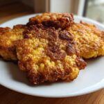 Four golden brown, crispy fried cornbread patties stacked on a white plate.