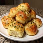 A stack of golden brown, glistening garlic breadsticks topped with herbs and Parmesan cheese on a white plate.