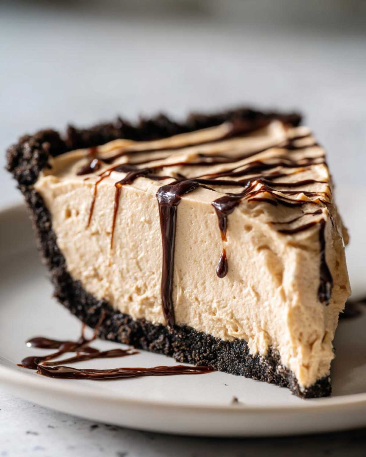 A close-up of a decadent slice of peanut butter pie with a dark crust and chocolate syrup drizzled on top.