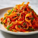 Close-up of a mound of vibrant raw carrot salad ribbons tossed in a spicy dressing and topped with sesame seeds.