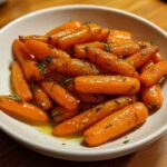A close-up of glossy, bright orange roasted baby carrots seasoned with herbs and sitting in a light glaze in a white bowl.